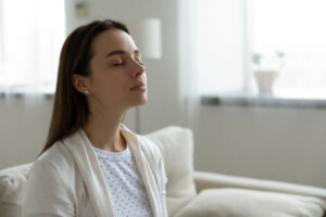 Calm serene woman resting sitting on couch in modern living room closed her eyes breath fresh humidified air. Fatigue relief repose, boost inner balance and mindfulness, meditation practice concept breathing clean indoor air