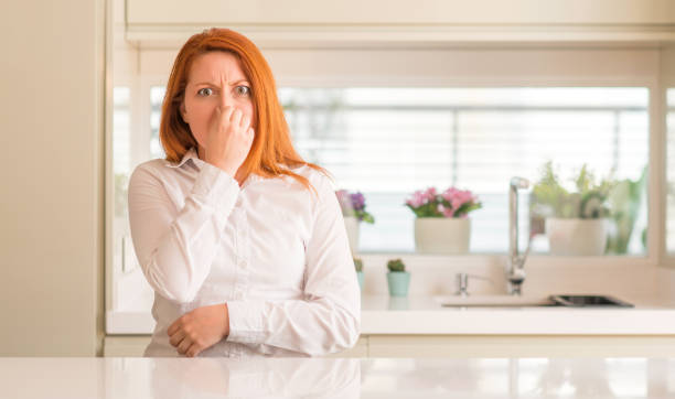 Redhead woman at kitchen smelling something stinky and disgusting, intolerable odors, holding breath with fingers on nose. Bad smell concept.